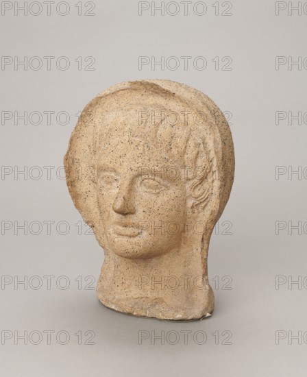 Votive head, Head of a young man covered with a shawl, from under which hair emerges in a fringe and falls in curls below the temples, covering the ears. Large, wide-open, convex eyes; large, straight nose; narrow mouth with wide, slightly curled, pouting lips. Face with a strong chin alluding to classical traditions. 4th century BC, Central Italy. Votive offering. Height 27.3 cm, width 20.5 cm, depth 18.5 cm, weight 3.5 kg., A sculpted head made of a speckled beige material sits against a plain light gray background, showing a stylized face with softly defined eyes, a straight nose, and closed lips, framed by a helmetlike or hooded shape with subtle carved hair details along the sides; the surface has a grainy texture with darker speckles, and the base appears slightly uneven and worn.