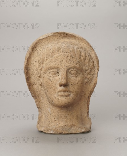 Votive head, Head of a young man covered with a shawl, from under which hair emerges in the form of a fringe and falls in curls below the temples, covering the ears. Large, wide-open, convex eyes; large, straight nose; narrow mouth with wide, slightly curled, pouting lips. Face with a strong chin alluding to classical traditions. 4th century BC, Central Italy. Votive offering. Height 27.3 cm; width 20.5 cm; depth 18.5 cm; weight 3.5 kg., A weathered sculpted head of a human with a hood-like surround, showing a neutral facial expression with closed lips and softly defined eyes and nose; the surface is textured and pitted, and the piece sits on a flat base. The visible colors are beige, tan, light brown, gray, and small darker speckles.