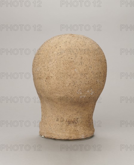 Votive head, Head of a young man covered with a shawl, from under which hair emerges in the form of a fringe and falls in curls below the temples, covering the ears. Large, wide-open, and convex eyes, large, straight nose, narrow mouth with wide, slightly curled, as if pouting lips. The face with a strong chin alludes to classical traditions. 4th century BC, Central Italy, votive offering. Height 27.3 cm, width 20.5 cm, depth 18.5 cm, weight 3.5 kg., A roughly spherical object with a narrower cylindrical base sits centered against a plain background, showing a coarse, textured surface that appears grainy and uneven; the object is primarily tan with darker brown speckles and small patches of white, while the background and surface it rests on are shades of light gray.