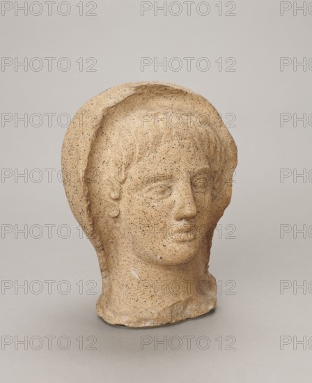 Votive head, Head of a young man covered with a shawl, from under which hair emerges in the form of a fringe and falls in curls below the temples, covering the ears. Large, wide-open, convex eyes; large, straight nose; narrow mouth with wide, slightly curled, as if pouting lips. Face with a strong chin alluding to classical traditions. 4th century BC, Central Italy. Votive offering. Height 27.3 cm, width 20.5 cm, depth 18.5 cm, weight 3.5 kg., A sculpted head of a human figure rendered in a sandy beige stone sits against a neutral light gray background, showing textured surface speckled with darker flecks; the face has defined eyes, nose, and closed lips with smooth contours, and short stylized hair framing the brow; a rounded hood or cap-like form encircles the head, creating a rim above the forehead and around the sides; the base of the neck is rougher and uneven where the sculpture was separated from a larger piece; overall colors visible are beige, light tan, darker brown flecks, a...