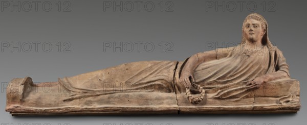 Sarcophagus lid, The deceased is depicted in a semi-reclining position, resting his left forearm on a pillow and holding a laurel wreath in his right hand. He wears a short-sleeved tunic and a cloak which, when thrown over the head, reveals the torso and left arm from shoulder to elbow; the right foot is visible from under the cloak. The human figure has very elongated proportions, with particularly strikingly long legs relative to the torso. 2nd half of the 2nd century BC, Etruria. Sarcophagus; height 71 cm, width 203 cm, depth 48 cm, weight 108 kg., A long horizontal terracotta sculpture of a reclining human figure on a low rectangular base, shown in profile and three-quarter view, with a draped garment creating flowing folds across the torso and legs; the figure rests one arm along the side and the other bent with the hand holding a wreath-like object, and the head is slightly raised with short textured hair and a serene facial expression; the surface shows cracks and patched joins,...