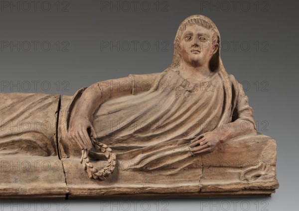 Sarcophagus lid;, The deceased is shown in a semi-reclining position, resting his left forearm on a pillow and holding a laurel wreath in his right hand. He wears a short-sleeved tunic and a cloak which, when thrown over the head, reveals the torso and left arm from shoulder to elbow; the right foot is visible from under the cloak. The human figure has very elongated proportions, with particularly disproportionate legs. Date: 2nd half of the 2nd century BC. Place of origin: Etruria. Object type: sarcophagus. Dimensions: height 71 cm, width 203 cm, depth 48 cm; weight 108 kg., A reclining stone figure with a pale beige and light brown surface wears draped garments with visible folds; the figure's head is covered by a hood-like drapery and shows a neutral facial expression with carved eyes and short textured hair. One arm rests along the side while the other hand holds a wreath of carved leaves, both hands and forearms showing similar beige and brown tones with subtle reddish patches. Th...