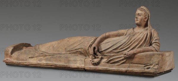 Sarcophagus lid, The deceased in a semi-reclining position, resting his left forearm on a pillow and holding a laurel wreath in his right hand. Dressed in a short-sleeved tunic and a cloak, which when thrown over the head reveals the torso and left arm from shoulder to elbow. The right foot is visible from under the cloak. Proportions of the human figure are very elongated, with especially long legs in disproportion to the torso. 2nd half of the 2nd century BC, Etruria. Sarcophagus; height 71 cm, width 203 cm, depth 48 cm, weight 108 kg., A reclining terracotta figure rendered in shades of beige, tan, brown, and hints of ochre rests on a rectangular base of similar earthy colors, showing the drape of a long garment with flowing folds across the body and arm. The figure lies on its side with a head turned upward, short stylized hair, and a cloth or veil falling behind it; a relaxed arm supports the upper body while the other hand holds a circular wreath-like object. The surface shows we...