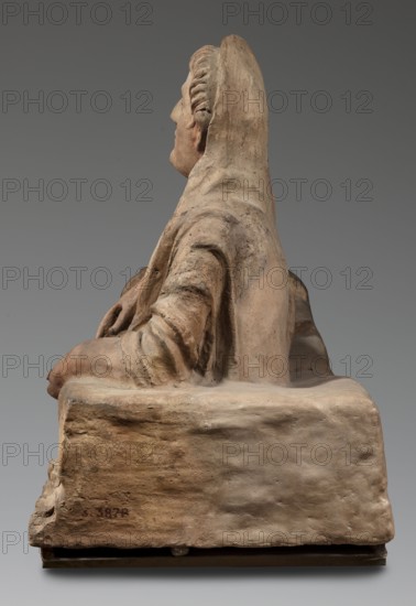 Sarcophagus lid, The deceased is shown in a semi-reclining position, resting his left forearm on a pillow and holding a laurel wreath in his right hand. He wears a short-sleeved tunic and a cloak which, when thrown over the head, reveals the torso and left arm from shoulder to elbow; the right foot is visible from under the cloak. The human figure has very elongated proportions, with notably long legs in disproportion to the torso. 2nd half of the 2nd century BC, Etruria. Sarcophagus; height 71 cm, width 203 cm, depth 48 cm, weight 108 kg., A side view of a seated sculptural figure carved from a pale, beige-toned material, showing the figure draped in a hooded garment with visible folds and textured surface; the figure's face is in profile with a slightly darker skin tone than the garment, and a hand rests on the lap emerging from a rolled sleeve; the base supporting the figure is rough-textured with earthy beige and light brown variations, and the background is a smooth gradient of gr...