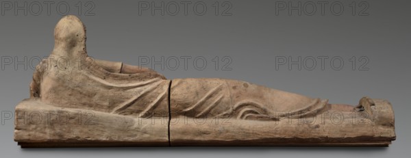 Sarcophagus lid, The deceased is depicted semi-reclining, resting his left forearm on a pillow and holding a laurel wreath in his right hand. He wears a short-sleeved tunic and a cloak which, when thrown over the head, reveals the torso and left arm from shoulder to elbow; the right foot is visible beneath the cloak. The human figure has very elongated proportions, with notably long legs in disproportion to the torso. Date: 2nd half of the 2nd century BC. Origin: Etruria. Object type: sarcophagus. Dimensions: height 71 cm, width 203 cm, depth 48 cm; weight 108 kg., A reclining sculpture of a human figure viewed from the side, made of a weathered clay or stone material in beige and light brown tones, lying on a rectangular base that shares similar beige and brown coloration; the figure is draped in a textured garment with visible folds and creases, showing dirt and darker brown spots in places, and the head is covered or smoothed without facial detail; there is a visible break where two...