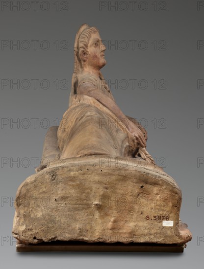 Sarcophagus lid, The deceased is depicted in a semi-reclining position, resting his left forearm on a pillow and holding a laurel wreath in his right hand. He wears a short-sleeved tunic and a cloak which, when thrown over the head, reveals the torso and left arm from shoulder to elbow; the right foot is visible from under the cloak. The human figure has very elongated proportions, with notably long legs in relation to the torso. 2nd half of the 2nd century BC, Etruria. Sarcophagus; height 71 cm, width 203 cm, depth 48 cm, weight 108 kg., A reclining sculptural figure viewed from the foot, showing a weathered stone or terracotta surface in shades of beige, tan, brown, and muted reddish tones; the figure has a visible head turned to the side with facial features and stylized hair, an upper torso and arms resting along the body, and hands clasped over the front; the draped form of a garment or covering extends along the length of the figure, with textured, worn edges and small pits and h...