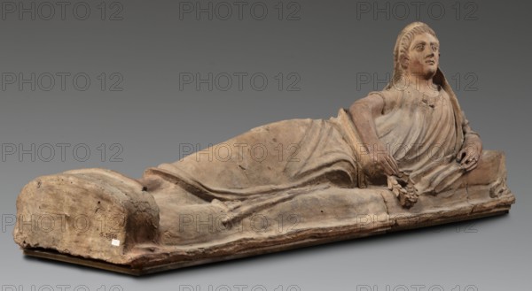 Sarcophagus lid, The deceased is shown in a semi-reclining position, resting his left forearm on a pillow and holding a laurel wreath in his right hand. He wears a short-sleeved tunic and a cloak which, when thrown over the head, reveals the torso and left arm from shoulder to elbow; the right foot is visible from under the cloak. The human figure has very elongated proportions, with notably disproportionate legs. 2nd half of the 2nd century BC, Etruria. Sarcophagus; height 71 cm, width 203 cm, depth 48 cm, weight 108 kg., A reclining terracotta figure on a rectangular base, shown in profile with a draped garment and a hooded head, displaying a few areas of smooth and rough texture; the figure's visible hand holds a small cluster of rounded forms while the other hand rests near the hip; the face has stylized features with wide eyes and a neutral expression; the surface shows worn and chipped areas and subtle contouring of fabric folds; colors visible include earthy terracotta tones, mu...