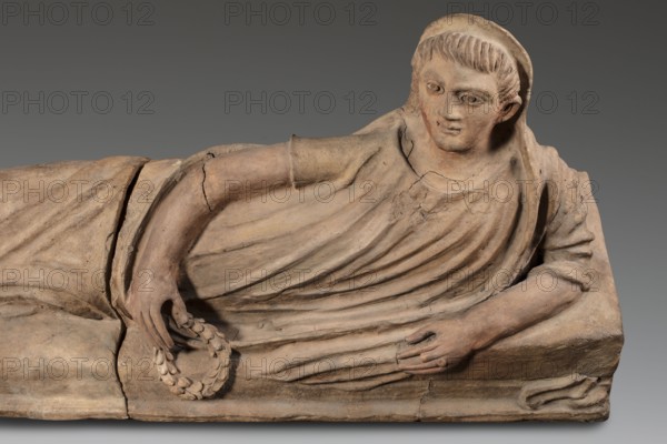 Sarcophagus lid, The deceased is depicted in a semi-reclining position, resting his left forearm on a pillow and holding a laurel wreath in his right hand. He wears a short-sleeved tunic and a cloak which, when thrown over the head, reveals the torso and left arm from shoulder to elbow; the right foot is visible from under the cloak. The human figure has very elongated proportions, with notably long legs in disproportion to the torso. 2nd half of the 2nd century BC, Etruria. Sarcophagus; height 71 cm, width 203 cm, depth 48 cm, weight 108 kg., A carved reclining figure rendered in beige and light brown tones rests on a rectangular base of similar color, wearing draped garments with visible folds and cracks; the figure's head is turned slightly with short carved hair and a rounded head covering, the facial features are softly modeled with eyes, nose, and lips, and one arm is bent to hold a circular laurel-like wreath while the other arm rests along the base, with the surface showing age...