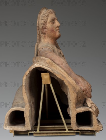 Sarcophagus lid, in a semi-reclining position. The deceased rests his left forearm on a pillow and holds a laurel wreath in his right hand. The figure is dressed in a short-sleeved tunic and a cloak, which, when thrown over the head, reveals the torso and left arm from shoulder to elbow. The right foot is visible from under the cloak. The proportions of the human figure are very elongated, with the legs being particularly striking in their disproportion to the torso. 2nd half of the 2nd century BC, Etruria; sarcophagus; height 71 cm, width 203 cm, depth 48 cm, weight 108 kg., A sculptural object in earth tones of tan, brown, and reddish-beige is shown against a gray background; it depicts a reclining human figure with a detailed head and visible facial features, a long textured hairstyle, and an extended arm resting along a curved form; the surface shows worn and cracked areas and variations of color from pale beige to darker brown and red; the base of the piece consists of hollow curv...