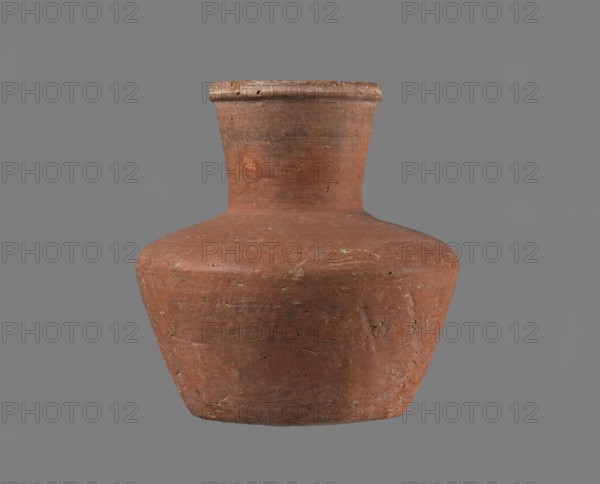 Jug, Small handleless jug with a biconical body, widest at about half its height; broad, rather flat shoulders. Neck wide, almost cylindrical, widening slightly toward a profiled, irregular spout that slopes down on one side and extends to form a small beak; spout cross-section triangular with an approx. 1 mm central indentation on the outer profile. Neck was formed separately and attached to the shoulders. Bottom almost flat, very slightly rounded. Made of Nile clay NB1/2? Vienna System. Outer surface and inner surface of rim and neck smoothed and covered with an engobe; remaining inner surface leveled. Red-brown color. Almost complete—small rim fragment missing; outer surface slightly worn in places. Old Kingdom, possibly 4th–6th Dynasty; provenance: Edfu. Vessel; height 11.4 cm; diameters: rim 5.5 cm, shoulders 10.8 cm, bottom 7.1 cm., A single rounded clay vessel with a narrow neck and flared rim sits against a plain background; the vessel has an uneven, slightly rough surface with...
