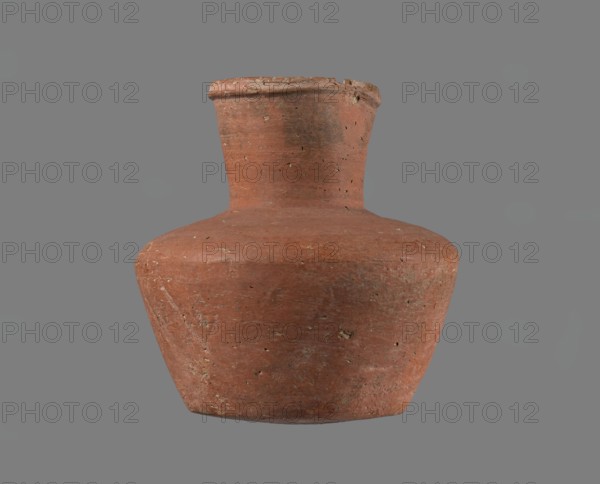 Jug. Small handleless jug with biconical body, widest at mid-height; broad, almost flat shoulders; separate neck glued to shoulders, almost cylindrical and widening slightly toward a profiled, irregular spout that slopes down on one side and forms a small projecting beak spout cross-section triangular with ~1 mm central indentation on outer profile. Bottom almost flat, very slightly rounded. Made of Nile clay NB1/2 ? — Vienna System. Outer surface and inner rim/neck leveled and coated with engobe and polished; remaining inner surface leveled. Red-brown. Nearly complete small fragment of rim missing; outer surface slightly worn. Attributed to Old Kingdom, possibly 4th–6th Dynasty; provenance: Edfu. Dimensions: height 11.4 cm; rim dia. 5.5 cm; shoulder dia. 10.8 cm; bottom dia. 7.1 cm., A weathered clay vessel with a rounded body and a narrow neck sits against a plain gray background; the pot shows a rough, textured surface with small pits and flecks, and its color is predominantly reddi...