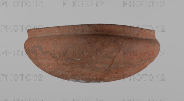 Medum-type bowl, ceramics; Egyptian pottery; clay vessel. Bowl with a hemispherical shape: walls bend gently around about 2/3 of the height of the vessel; straight rim perpendicular to the shoulders, slightly irregular; rounded bottom. Surface even; red color. Vessel glued together from several fragments. c. 2686–c. 2181 BC, Old Kingdom, Edfu. Vessel; height 5.5 cm; diameter 12.6 cm rim; diameter 13.5 cm shoulders., A rounded clay bowl shown against a plain gray background, with an overall reddish-brown color and areas of darker brown and faint black speckling; the surface appears rough and weathered with visible cracks and small chips, and there are traces of lighter tan and gray discoloration in spots.