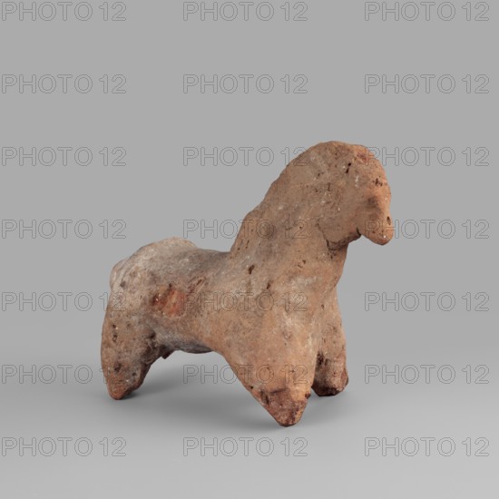 Horse figurine, horses; 664–30 BC Late Period–Ptolemaic Period; found at Edfu; figurine; height 8 cm, width 10 cm., A small, weathered clay figure shaped like an animal with a rounded head and short neck standing on a broad body with four stubby legs, showing a rough, pitted surface and irregular contours; the figure displays a palette of earthy browns and reddish-brown tones with patches of lighter beige and small darker spots, set against a smooth neutral gray background.
