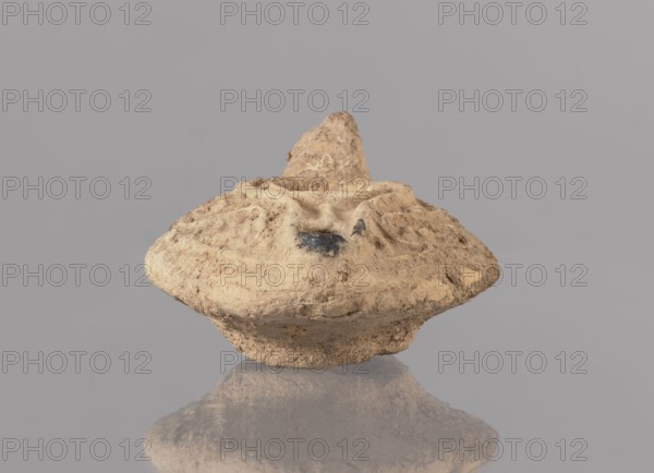 Oil lamp, deposit provenance, Islamic art, Arabic culture, Arabic pottery, utilitarian pottery, everyday life in the Middle East, Egyptian pottery, oil lamp with traces of use, plant tendril decoration. Oval lamp with short burner channel surrounded by a shaft similar to the disc. Large oil hole occupying the entire surface of the disc. Arms decorated with plant tendrils. Conical handle. Ring-shaped base with a star in the center. 2nd half of the 7th–8th century, Syro-Palestine; height 3.4 cm, width 11.0 cm, depth 8.0 cm., An irregular, roughly symmetrical object with a rounded, disk-like lower portion and a protruding, conical top sits centered against a flat, gray background; the object has a rough, textured surface in shades of beige, tan, and light brown with a few darker brown and black patches, and it casts a subtle reflection below on the glossy surface.