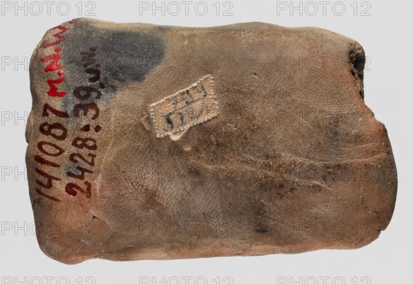 A mold for making an amulet lion, lions, casting mold, Ptolemaic Period 332–30 BC, found in Edfu; mold, height 7.1 cm, A roughly rectangular, worn object with an uneven, textured surface in shades of brown, tan, and beige, showing areas of darker brown and grayish-black discoloration; a small frayed textile patch in light beige with traces of dark pigment is affixed near the center; the edges are rounded and irregular with a small chipped area on one side; the surface displays rubbed, rough patterns and subtle linear impressions.