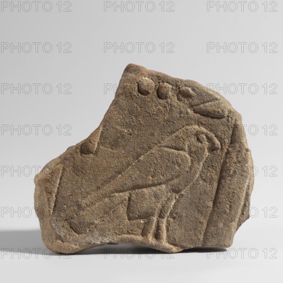 The Hor Hieroglyph - relief fragment, Horus mythology. Relief fragment with several preserved hieroglyphic signs. The preserved fragment features a schematic representation of a falcon the Hor sign shown in right profile, above which three ball-like signs are preserved, next to which a triangular sign is preserved. Sections of the side frames remain. 3rd–1st century BC; Ptolemaic Period, Edfu. Inscription, bas-relief. Height 17 cm, width 16 cm, depth 3 cm., A roughly triangular fragment of brown stone with a textured, weathered surface shows a carved outline of a bird in profile perched on a short base, with carved lines suggesting a beak, eye area, wings, tail, and legs; a few rounded carved depressions appear near the top edge and several linear incised shapes run along the right side; the stone displays shades of brown and tan against a pale gray background.