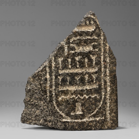Fragment of a block with the Sheshonq cartouche, Sheshonq ruler of Egypt; ca. 945–715 BC. Cartouches were processed loops of double-wound rope signifying protection; this relief fragment shows the cartouche of Pharaoh Sheshonq of the 22nd Dynasty. It is unknown whether this refers to the dynasty founder the biblical Shishak or to one of four other rulers of that name. Royal cartouches and hieroglyphic texts could be written vertically or horizontally; the characters inside this cartouche point to the left, indicating the pharaoh's figure faced the same direction. ca. 945–ca. 735 BC; Third Intermediate Period, 22nd Dynasty, Egypt. Bas-relief; height 15.5 cm, width 13 cm, depth 6 cm, weight 1.138 kg., A rough triangular stone fragment sits on a neutral surface against a gray background, showing a carved oval design with horizontal recessed bands and rounded edges; the stone's surface is textured and granular with a mix of dark gray, light gray, beige, and black tones, and a few lighter c...