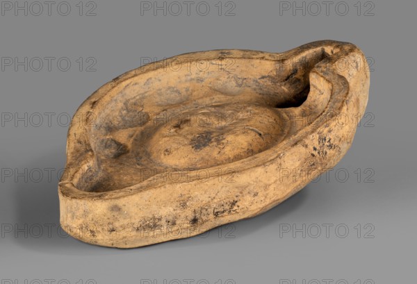 Lamp production mold, oil lamp ceramic mold, Roman lamps, material culture, 1st–2nd century, Roman Empire 27 BC–476, mold; height 4 cm, width 11.6 cm, depth 8.5 cm, A weathered shallow bowl-shaped object with an irregular oval outline, made of a rough textured material in shades of tan and brown with darker brown and blackened patches; its rim is uneven and slightly raised, curving inward to form a concave interior that has a central raised mound and shallow depressions, while one end shows a carved notch or spout-like indentation; the object rests on a smooth gray surface and casts a soft shadow.