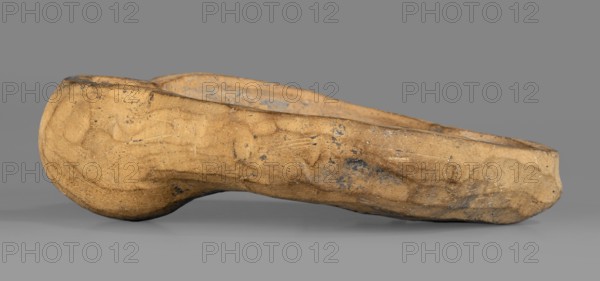 Lamp production mold, oil lamp mold of ceramic, Roman, 1st–2nd century Roman Empire, 27 BC–476 AD; mold; height 4 cm, width 11.6 cm, depth 8.5 cm., A roughly elongated, irregularly shaped object with a rounded end rests on a flat surface, showing a textured, worn surface with visible scuffs and small pits; the object displays primarily tan and beige tones with patches of darker brown and some grayish-blue discoloration, and the background and surface beneath are neutral gray.