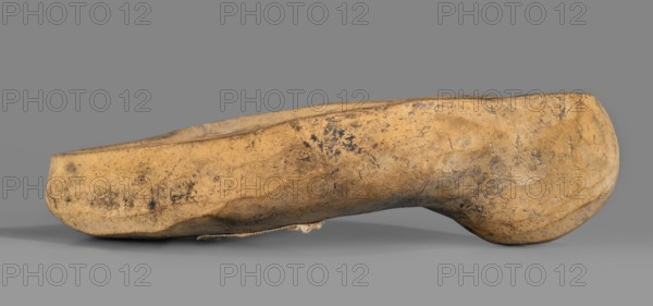 Lamp production mold, oil lamp ceramic mold, Roman lamps, 1st–2nd century, Roman Empire 27 BC–476, mold, height 4 cm, width 11.6 cm, depth 8.5 cm, The image shows a single worn shoe form or clog-like object lying on its side against a neutral gray background; the object has an overall beige to tan color with patches of darker brown and black discoloration and surface scuffs, a rounded heel area and a slightly flattened toe area, visible cracks and textured wear across its surface, and a small frayed piece of fabric or material protruding from the underside near the middle.
