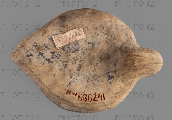 Lamp production mold, oil lamp ceramic mold, Roman, 1st–2nd century CE Roman Empire, 27 BC–476, mold; height 4 cm, width 11.6 cm, depth 8.5 cm, A roughly rounded, irregular object with a textured, worn surface showing shades of tan, beige, brown, gray, and black speckling; a few areas of darker gray-black patches contrast with lighter tan and beige regions. A small, pale fabric patch with frayed edges is attached near the upper center. The object has an asymmetrical protrusion on one side and subtle ridges and indentations along its edges. The background is a uniform medium gray.