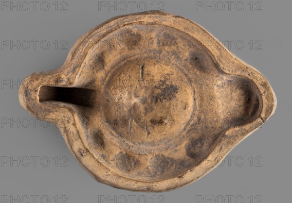 Lamp production mold, oil lamp ceramic mold, Roman lamps, material culture, Roman Empire, 1st–2nd century, mold; height 4 cm, width 11.6 cm, depth 8.5 cm, A worn, shallow ceramic object viewed from above with an overall tan and brown color, showing darker brown and blackened areas and patches of discoloration; it has an irregular rounded shape with a central circular depression, a wider shallow rim with several rounded indentations, and two protruding extensions at opposite sides—one forming a tapered spout-like opening and the other a rounded pour or recess; the surface shows textured roughness, scratches, and small chips, and the object rests on a flat gray background.