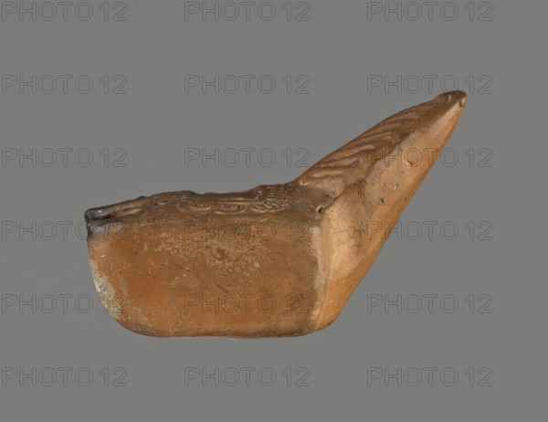 Oil lamp, unknown Pannonian workshop. Oil lamp with leaf ornament, Roman three-burner lamp. Made from a mold. Rectangular, flat vessel with a slightly recessed rectangular disc surrounded by a stylized egg-shaped ornament. Three olive holes placed at regular intervals within the disc. Leaf-shaped handle on the longer side. Opposite side has three short circular burners with large wick holes; all are charred. 1st–2nd century, Pannonia. Oil lamp; height 3.3 cm, width 7.3 cm, depth 5.1 cm., A single clay object photographed against a plain gray background, showing a worn rounded base and an elongated pointed extension; the surface is textured with irregularities and shallow decorative impressions, and there are rough patches and small pitted areas. The visible colors include shades of brown, orange-brown, and tan, along with some darker brown and grayish spots.