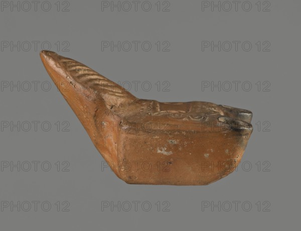 Oil lamp, unknown Pannonian workshop. Oil lamp with leaf ornament, Roman three-burner lamp. Mold-made rectangular, flat vessel with a slightly recessed rectangular disc surrounded by stylized egg-shaped ornament. Three olive holes placed at regular intervals within the disc. Leaf-shaped handle on the longer side; opposite side with three short circular burners with large wick holes, all charred. 1st–2nd century, Pannonia. Height 3.3 cm, width 7.3 cm, depth 5.1 cm., A small sculpted object sits against a plain gray background, appearing to be made of a brownish-orange clay with areas of darker brown and blackened discoloration; the form has an elongated, tapered projection with carved diagonal grooves on its top surface and a broader, more rectangular base with a gently curved front edge; the surface shows rough texture with pitting and small patches of lighter and darker tones, and the piece displays some worn and smoothed areas along edges and raised details.