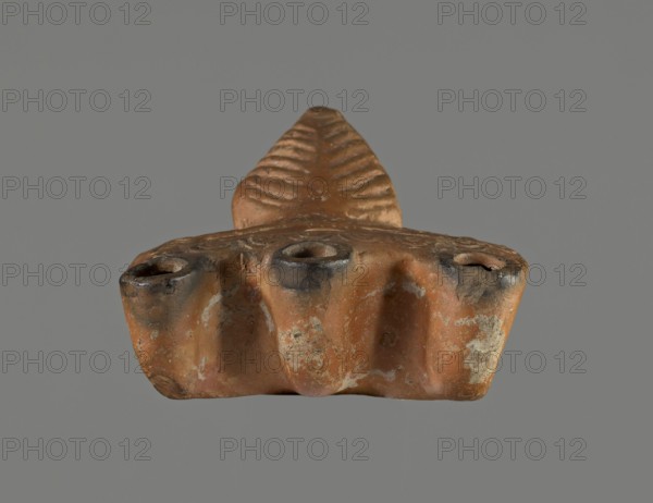 Oil lamp, Pannonian workshop. Oil lamp with leaf ornament, Roman three-burner lamp. Mold-made, rectangular flat vessel with slightly recessed rectangular disc surrounded by stylized egg-shaped ornament. Three olive holes placed at regular intervals within the disc. Leaf-shaped handle on the longer side; opposite side with three short circular burners with large charred wick holes. 1st–2nd century. Height 3.3 cm; width 7.3 cm; depth 5.1 cm., A small sculptural object with a worn, textured surface in shades of brown, orange, black, gray, and pale beige is shown against a neutral gray background; the object has a triangular, ridged projection rising from a broader base that features three rounded depressions or openings along its front edge, darkened around the rims, and the surface shows patches of abrasion and lighter mineral-like deposits.