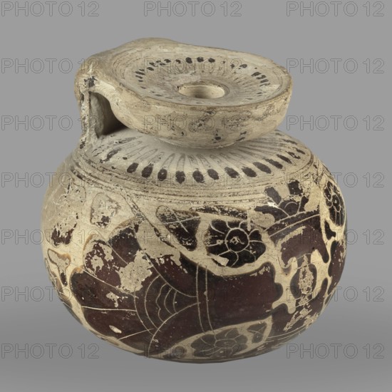 Orientalizing aryballos with depictions of lotus flowers;, repossession Silesia; lotus iconography; vessel for oils and ointments; Greek pottery, 580–570 BC; Corinth; vessel aryballos; height 6.8 cm; width 7.4 cm; weight 0.112 kg, A round ceramic vessel with a rounded body and a short neck topped by a flat, slightly concave lid-like rim connected by a short spout-like element, shown against a solid gray background. The surface shows a worn pale beige base color with areas of darker brown and black painted decoration forming floral shapes and abstract motifs, along with lighter areas where the surface is chipped. The rim and shoulder area display a pattern of short dark strokes around the opening, and the vessel surface has visible scratches, chips, and uneven texture indicating wear. The overall color palette includes beige, dark brown, black, and hints of off-white from exposed ceramic.