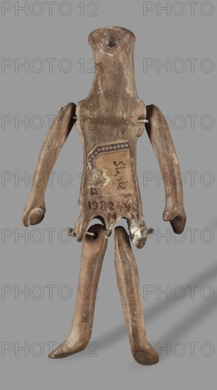 Doll with movable limbs, unknown Corinthian workshop author ?, restitution provenance, toys, dolls, human figurines, 500–450 BC, Corinth place of origin ?, terracotta figurine, height 13.9 cm, width 4.1 cm, depth 1.9 cm, weight 0.05 kg, A small humanoid figure made of a brown, weathered material stands against a plain gray background; the figure has an elongated, flattened head and torso with a rough, uneven surface showing lighter and darker brown patches and areas of wear, a few small white flecks, and some smoother, shinier spots. The figure has movable arms and legs attached with short metal rods or wires, the arms are curved and taper to rounded ends while the legs are long and slightly bent with rounded feet. There is a small rectangular patch of a darker tan color on the torso with a decorative border in a darker brown, and the lower torso shows some irregular lighter-colored remnants where the material appears chipped or worn. A faint shadow falls to the right and below the fig...