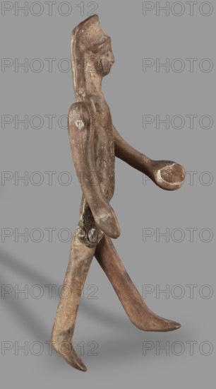 Doll with movable limbs, unknown Corinthian workshop, restitution provenance, toys, dolls, human figurines, 500–450 BC, Corinth, figurine; height 13.9 cm, width 4.1 cm, depth 1.9 cm, weight 0.05 kg, A worn carved wooden figure shown in profile against a plain gray background, with a smooth elongated head and stylized facial features, a long neck, and a narrow torso; the figure has articulated limbs with one arm extended forward ending in a rounded hand and the other arm resting alongside the body, and legs positioned as if walking with one foot pointed forward and the other angled back. The surface shows weathered textures, scuffs, and areas of lighter residue, with overall brown tones ranging from dark brown to lighter tan and small patches of off-white.