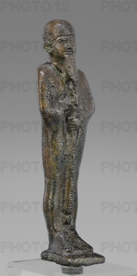Figurine of the god Ptah; creator, restitution provenance; Ptah mythology; Egyptian deity; ankh and wa's scepter iconography; beard facial hair; Egyptian art, Late Period, 664–332 BC; material/function: figurine; dimensions: height 14.7 cm, width 4 cm, depth 3 cm; weight 0.272 kg., A narrow upright statuette of a standing human figure with a long beard and a close-fitting cap, rendered in a textured metallic surface showing a mix of dark brown, greenish patina, and some muted yellowish tones, arms folded or held close to the torso, a long robe that falls straight to a flat base, subtle surface pitting and irregularities across the figure, and a neutral gray gradient background.