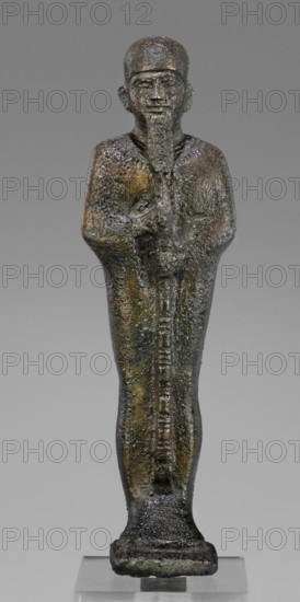 Figurine of the god Ptah; maker, restitution provenance; Ptah mythology; Egyptian deity; ankh and wa's scepter iconography; beard facial hair; Egyptian art; Late Period, 664–332 BC, Egypt; figurine; height 14.7 cm, width 4 cm, depth 3 cm, weight 0.272 kg., A weathered statuette of a standing human figure with a long beard and a rounded cap, rendered in a textured metal surface showing a mix of dark brown, green, and gray patina; the figure's arms are held close to the body and appear to clasp a vertical object that extends down the length of the torso to a rectangular base, with surface irregularities and areas of darker and lighter mottling throughout, all shown against a neutral gray background.