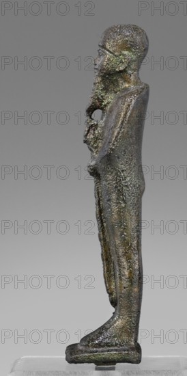 Figurine of the god Ptah; unknown creator; restitution provenance; Ptah mythology; Egyptian deity; ankh and wa's scepter iconography; beard; Egyptian art; Late Period, 664–332 BC; figurine; height 14.7 cm, width 4 cm, depth 3 cm, weight 0.272 kg., A slender vertical metal figure with a textured, rough surface in shades of dark brown, greenish patina, and hints of black, shown in profile standing on a small rectangular base; the head is rounded with a defined nose and chin, a long textured beard extends along the chest, and the body tapers down to feet that rest flat on the base; the figure wears a long, close-fitting garment with surface irregularities and a few areas of lighter green and brown sheen, and there is a visible hollow or gap near the midsection.