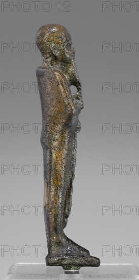 Figurine of the god Ptah; creator, restitution provenance; Ptah mythology; Egyptian deity; ankh iconography; wa's scepter iconography; beard facial hair; Egyptian art, 664–332 BC, Late Period, Egypt; figurine; height 14.7 cm, width 4 cm, depth 3 cm, weight 0.272 kg., A small bronze statuette of a standing human figure shown in profile against a gray background, with a textured surface showing areas of green and dark brown patina and patches of lighter brown and yellowish tones; the figure has a rounded head, visible facial features in profile, and a long beard or facial extension; the body is elongated and wrapped by a cloak or garment that runs straight down the back and sides, with the arms held close to the torso and hands meeting at the midline; the figure stands on a flat rectangular base that has darker green corrosion at its lower edge.