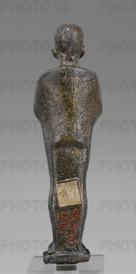 Figurine of the god Ptah; unknown creator; restitution provenance; Ptah mythology; Egyptian deity; ankh and wa's scepter iconography; beard facial hair; Egyptian art, Late Period, 664–332 BC; Egypt; figurine; height 14.7 cm, width 4 cm, depth 3 cm, weight 0.272 kg., A small upright figurine shown from the rear, with a rounded head, visible ears, and a tapered body standing on a rectangular base. The surface is textured and pitted with an uneven, aged patina. Colors visible include dark brown, greenish areas, and patches of lighter brown or ochre on the back and head, with a few darker gray and black tones across the figure. The figure is positioned against a neutral gray background and rests on a clear display stand.
