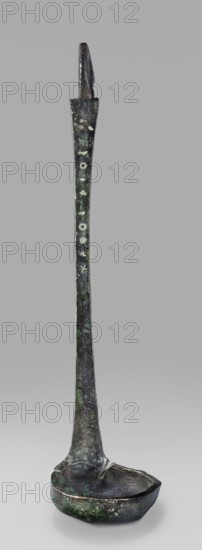 Ladle with a handle in the form of a goose's head. Ladle with a shallow bowl ending in a wide, flat rim. The flat handle, attached perpendicularly, ends in a hooked bend in the shape of a goose's head. Engraved Greek inscription on the inside. 5th century BC, Greece. Ladle; height 27 cm, width 6 cm, weight 0.12 kg., A tall slender metal object with a long narrow handle that widens slightly toward the base, standing upright against a plain gray background; the handle and bowl show a weathered, uneven surface with patina in shades of dark brown, green, and dull gray, and a few lighter green spots; at the top the handle ends in a hooked or notched tip, and the bottom flares into a shallow, rounded bowl with an irregular rim and a rough, pitted texture.