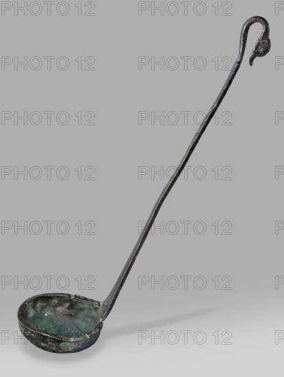 Ladle with a handle in the form of a goose's head. Ladle with a shallow bowl ending in a wide, flat rim. The flat handle, attached perpendicularly, ends in a hooked bend in the shape of a goose's head. Engraved inscription on the inside. 5th century BC, Greece. Ladle; height 27 cm, width 6 cm, weight 0.12 kg. Greek inscriptions; Zeus myth.., An aged metal ladle with a long, slightly curved handle ending in a hooked top sits against a plain gray background; the bowl is shallow and round with uneven, dented edges and a patina showing green, dark brown, and black tones, while the handle shows worn dark metallic colors with hints of greenish corrosion and small lighter spots where the surface is abraded.