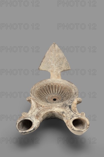Two-burner lamp decorated with a rosette motif, oil lamp, end of the 1st century, Asia Minor; height 10.2 cm, width 21.5 cm, depth 13.5 cm, weight 0.260 kg., An off-white and beige ceramic object with areas of darker gray and black discoloration, shaped with a central shallow bowl featuring radiating fluted ridges and a small central hole, a triangular vertical backplate rising from the rear with faint carved floral-like motif, and two rounded protruding spouts at the front connected by a curved bridge, all resting against a plain mid-gray background.