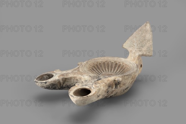 Two-burner lamp decorated with a rosette motif, two-burner oil lamp with rosettes iconography, end of the 1st century, Asia Minor; oil lamp; height 10.2 cm, width 21.5 cm, depth 13.5 cm, weight 0.260 kg, An off-white and beige ceramic oil lamp with areas of darker gray and black discoloration sits against a neutral gray background; the lamp has a shallow bowl with a radiating fluted pattern carved into its interior, two rounded spouts extending from the front edge, and a raised triangular fin at the rear decorated with a faint carved motif; the surface shows textured wear, small chips, and uneven coloration with spots of darker residue, and the overall shape is elongated and asymmetrical.