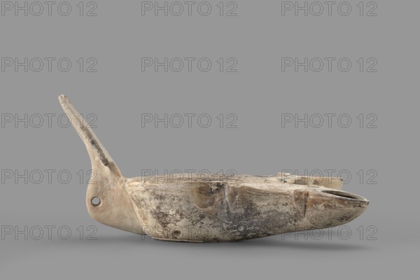 Two-burner lamp decorated with a rosette motif, two-burner oil lamp with rosettes iconography, end of the 1st century, Asia Minor; oil lamp; height 10.2 cm, width 21.5 cm, depth 13.5 cm; weight 0.260 kg., A weathered, shallow bowl-like object with an elongated pointed handle rests on a neutral gray surface against a gray background; the object shows an aged, mottled surface with tones of beige, off-white, tan, brown, and darker gray patches, a rounded body with a small circular hole near the base of the handle, a carved groove and subtle raised areas along the rim, and areas of surface wear and discoloration giving a rough, textured appearance.