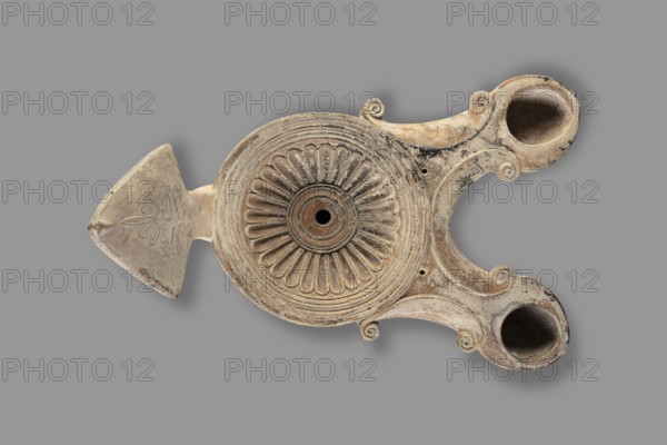 Two-burner oil lamp decorated with a rosette motif, end of the 1st century, Asia Minor; height 10.2 cm, width 21.5 cm, depth 13.5 cm, weight 0.260 kg., An off-white to beige ceramic object with areas of darker brown and black discoloration, shaped with a large central circular section decorated with a raised radial floral motif and concentric ridges, a pointed triangular projection on one side with incised petal-like ornament, and two rounded basin-like projections on the opposite side connected by curved, scroll-accented arms; the piece shows varied surface texture from smooth to eroded, with shadows suggesting hollow cavities in the basin areas, all set against a solid gray background.