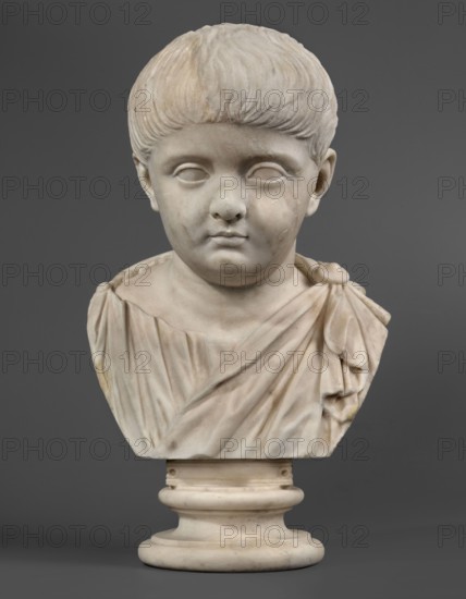 Portrait of a prince from the Julio-Claudian dynasty, unknown Roman workshop, boy portrait, Roman sculpture, antique bust, dated 40–70 CE, Roman Empire 27 BC–476, sculpture; height 36 cm, width 21 cm, depth 18 cm, weight 11.55 kg, A marble bust of a young child is shown against a gray background, carved with short textured hair and smooth facial features including closed or blanked eyes, a rounded nose, and a small mouth; the child wears a draped garment with folds and a shoulder knot, and the sculpture rests on a rounded cylindrical base. The visible colors are white and off-white tones in the marble with subtle beige and pale yellow discolorations, and a solid mid-gray background.