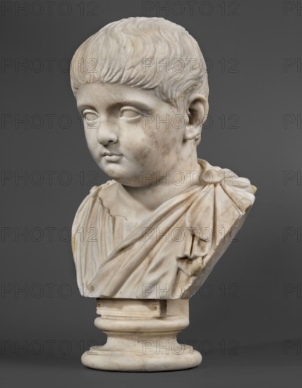 Portrait of a prince from the Julio-Claudian dynasty, unknown Roman workshop; Roman sculpture, antique bust, boy portrait; Roman Empire 27 BC–476, dated 40–70 CE; marble sculpture, height 36 cm, width 21 cm, depth 18 cm, weight 11.55 kg., A carved marble bust of a young child shown from the shoulders up on a rounded plinth, rendered in pale off-white and creamy beige tones with subtle veins and weathered spots, the child has a rounded face with short, textured hair, closed eyes lacking detailed pupils, a small nose and closed lips, and a soft, neutral expression; the figure wears a draped garment with folded, flowing folds and a gathered shoulder detail, the sculpture shows smooth and slightly worn surfaces with faint discolored patches, all set against a solid dark gray background.