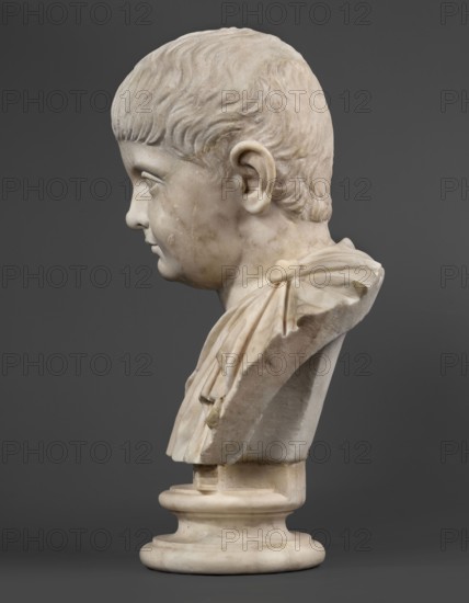 Portrait of a prince from the Julio-Claudian dynasty, unknown Roman workshop; boy portrait, Roman sculpture, antique bust, dated 40–70 CE, Roman Empire 27 BC–476; sculpture; height 36 cm, width 21 cm, depth 18 cm, weight 11.55 kg., A marble bust of a child shown in profile against a dark gray background, with pale off-white and creamy tones in the stone, subtle veins and weathering in beige and light brown, short carved hair, a rounded cheek and ear, a draped garment over the shoulder with carved folds, and the sculpture mounted on a circular base of similar pale stone.