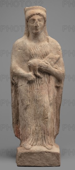 Figurine of a woman with a polos on her head, holding a piglet. Figurine of a woman standing on a rectangular base. The woman is dressed in a finely pleated chiton reaching down to her bare feet, with a semicircular neckline. A himation, draped over her shoulders like a shawl, covers her arms to the elbows. On her head she wears a cylindrical polos, slightly widening upwards and folded into a veil flowing down the back. Hair protrudes from beneath the headdress, flowing in wavy patterns to the sides. The face is full, with a rounded chin, large eyes, and fleshy lips drawn into a characteristic smile. In her left hand, bent at the elbow, the woman holds a piglet; with her right hand she supports the animal's hind leg. 5th century BC, Southern Italy Italy. Terracotta figurine; height 28 cm, width 10 cm, depth 10 cm, weight 0.57 kg. Pigs iconography; female figurines., A weathered standing statue of a robed figure carved from light brown stone, shown full-length against a plain gray backg...