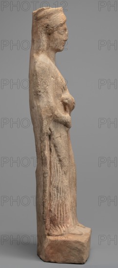 Figurine of a woman standing on a rectangular base. The woman is dressed in a finely pleated chiton reaching down to her bare feet, with a semicircular neckline. A himation, draped over her shoulders like a shawl, covers her arms to the elbows. On her head she wears a cylindrical polos, slightly widening upwards and folded into a veil flowing down the back. Hair protrudes from beneath the headdress, flowing in wavy patterns to the sides. The face is full, with a rounded chin, large eyes, and fleshy lips drawn into a characteristic smile. In her left hand, bent at the elbow, the woman holds a piglet; with her right hand she supports the animal's hind leg. Pigs iconography; female figurines. 5th century BC, Southern Italy Italy. Terracotta figurine; height 28 cm, width 10 cm, depth 10 cm, weight 0.57 kg., A weathered stone statue of a standing female figure shown in profile, with a tall headdress or hair pulled back, a visible nose and chin, and a calm facial expression; the figure's arm...