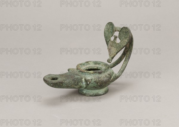 Oil lamp, 1st century, Pozzuoli, oil lamp, weight 0.524 kg, A small, aged oil lamp with a rounded body and a short spout sits on a flat surface; the lamp has a raised circular opening on top and a curved handle that rises and forms a decorative, heart-shaped loop at its end with some missing pieces. The surface of the lamp shows a rough, corroded texture with patches of green and darker green patina and areas of brownish and gray discoloration. The background and surface beneath the lamp are a smooth, pale beige-gray color.