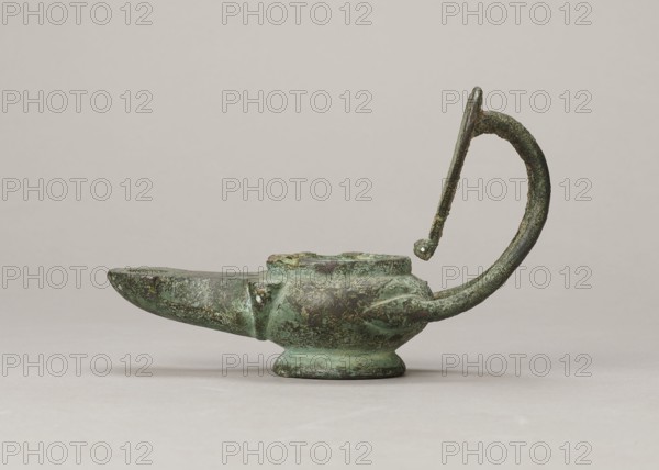 Oil lamp, maker, 1st century; Pozzuoli possibly; oil lamp; weight 0.524 kg., An aged metal lamp with a patina of green and dark brown sits on a neutral pale surface against a light beige background; the lamp has a rounded body on a small base, a tapered spout extending to the left, and a tall curved handle rising and arching back toward the body, all showing textured corrosion and mottled green and brown tones.